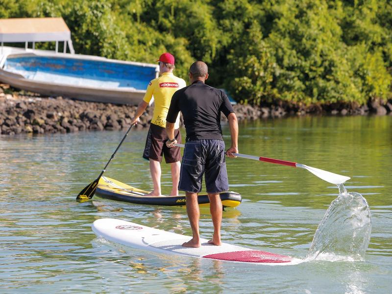 Zwei Männer stehen auf Paddleboards und paddeln auf ruhigem Wasser vor grünem Ufer.
