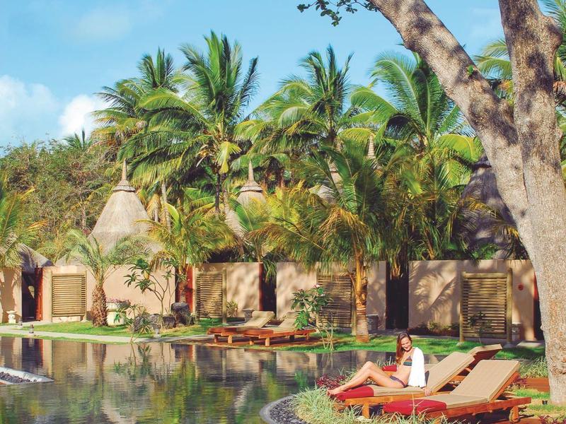 Ein Pool mit zwei roten Liegestühlen, umgeben von Palmen und Bäumen bei sonnigem Himmel.