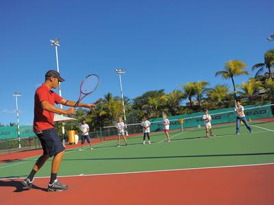 Person spielt Tennis auf einem sonnigen Außenplatz mit mehreren Zuschauern im Hintergrund.