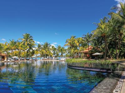 Large pool with clear blue water surrounded by palm trees and umbrellas under a blue sky.