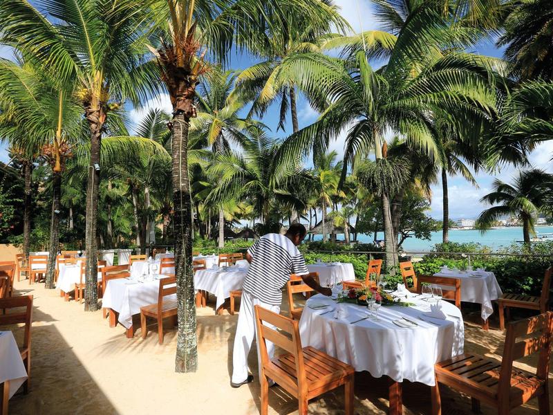 Beachside restaurant with white tablecloths, wooden chairs, and palm trees in the background.