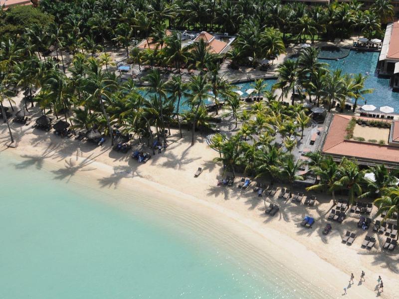Beach with turquoise water, palm trees, and shaded umbrellas next to a hotel pool.