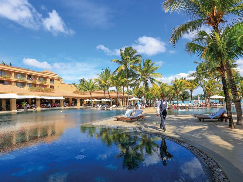Hotel with large pool, palm trees, and umbrellas under a blue sky.