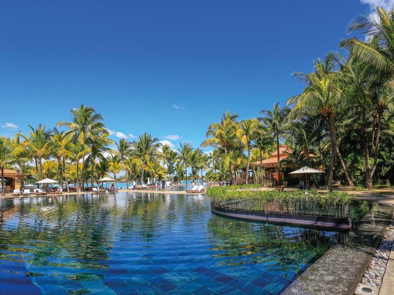 Large pool with clear blue water surrounded by palm trees and umbrellas under a blue sky.