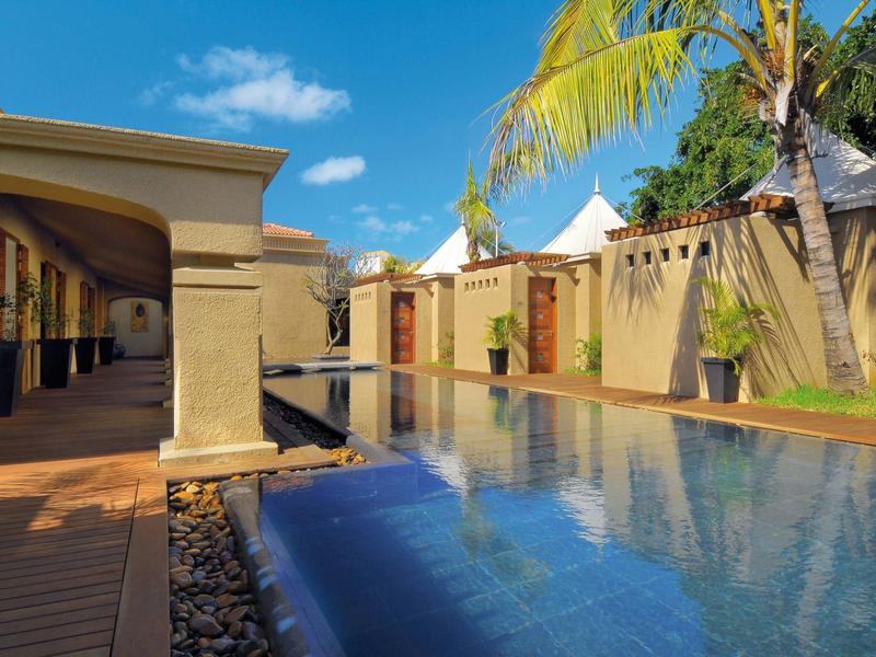 Modern hotel pool with blue water, sandstone walls, and palm trees under a clear blue sky.