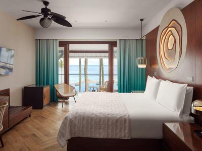 Helles Hotelzimmer mit großem Bett, Holzdekor, türkisfarbenen Vorhängen und Meerblick.