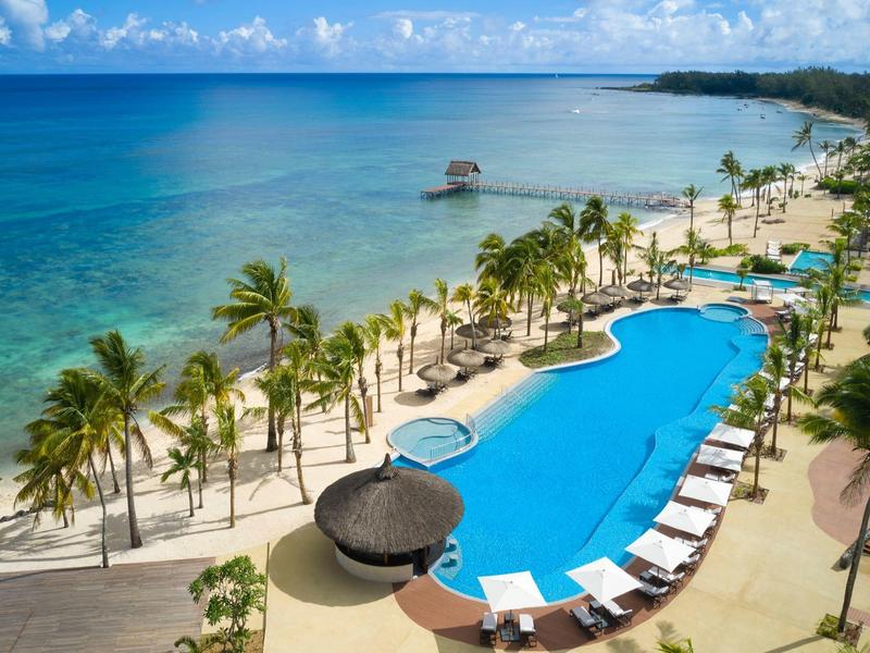 Lang zwembad met ligbedden en palmbomen naast een rustige blauwe oceaan onder een deels bewolkte lucht.