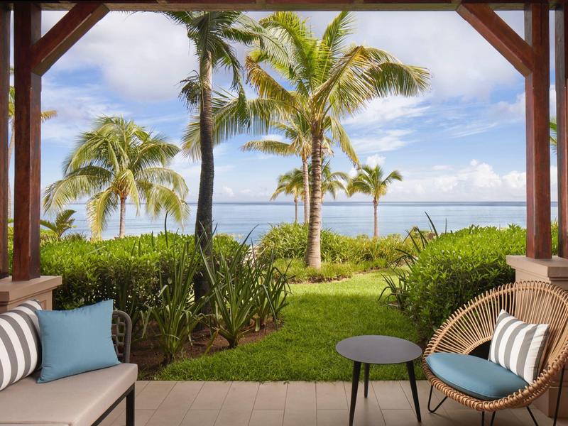 Uitzicht vanaf een overdekt terras op een tropische tuin met palmbomen en de zee op de achtergrond.