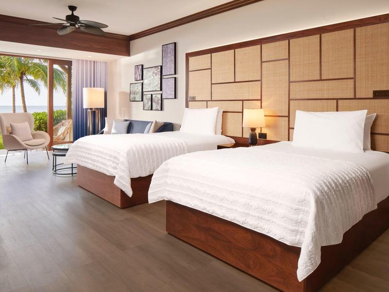 Lichte hotelkamer met twee bedden, gezellige zithoek en uitzicht op palmbomen en zee.