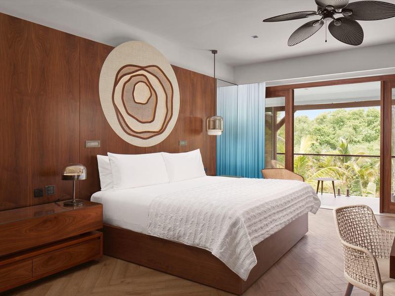 Modern hotelkamer met houten wanden, groot bed, plafondventilator en balkon met uitzicht op groen.