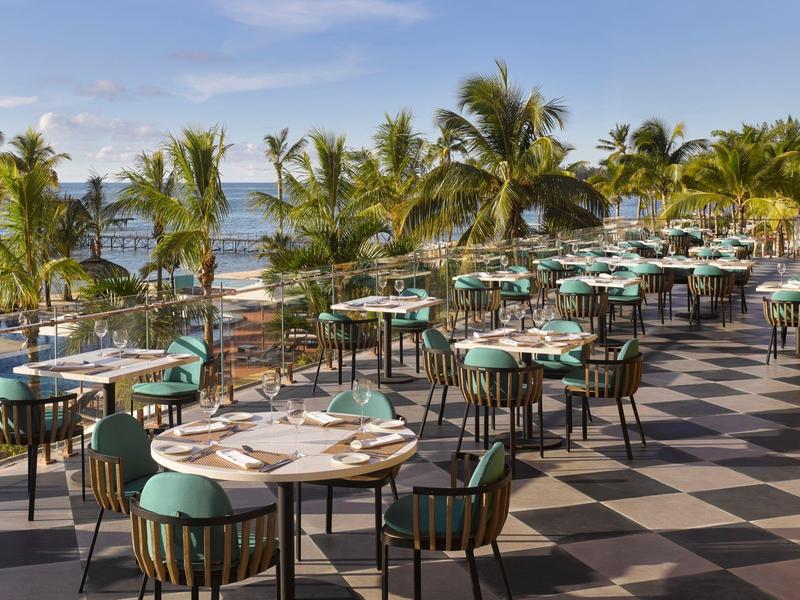 Terras van een restaurant met tafels en stoelen bij de zee en palmbomen op de achtergrond.