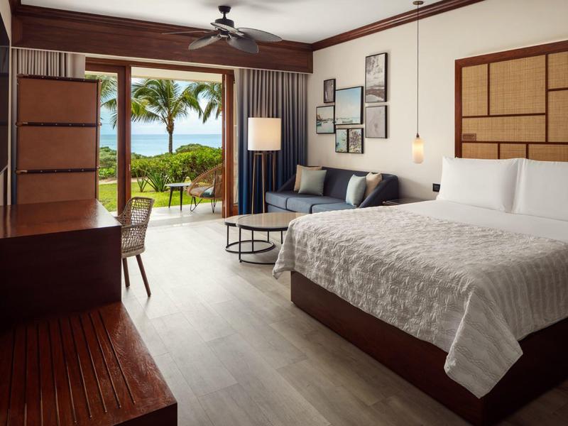 Lichte hotelkamer met tweepersoonsbed, zithoek en uitzicht op tropische planten en zee.