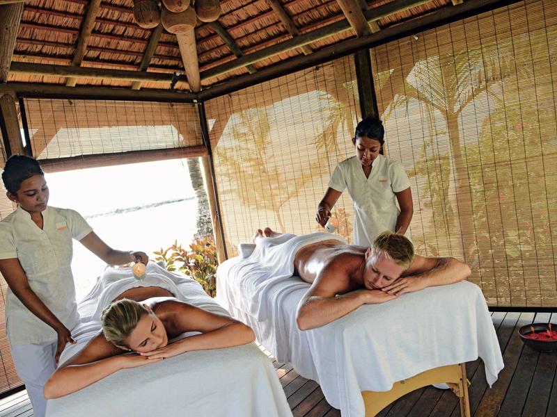 Zwei Personen genießen Massage in Holzhütte, umgeben von Bambusrollos und Dach aus Holzblättern.