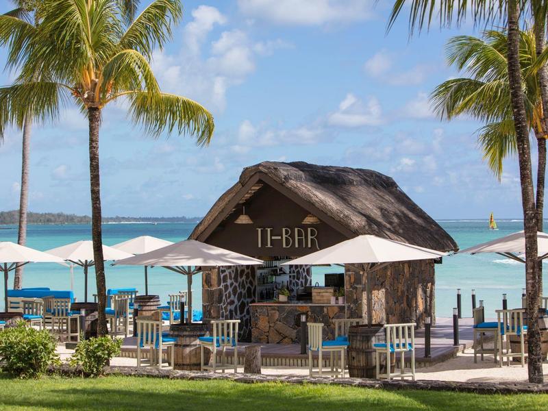 Strandbar mit Strohdach, Tischen, Sonnenschirmen, Palmen und blauem Meer im Hintergrund.