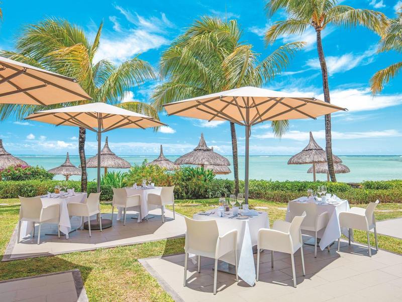 Strandrestaurant mit weißen Tischen, Stühlen und großen Sonnenschirmen vor Palmen und Meer.