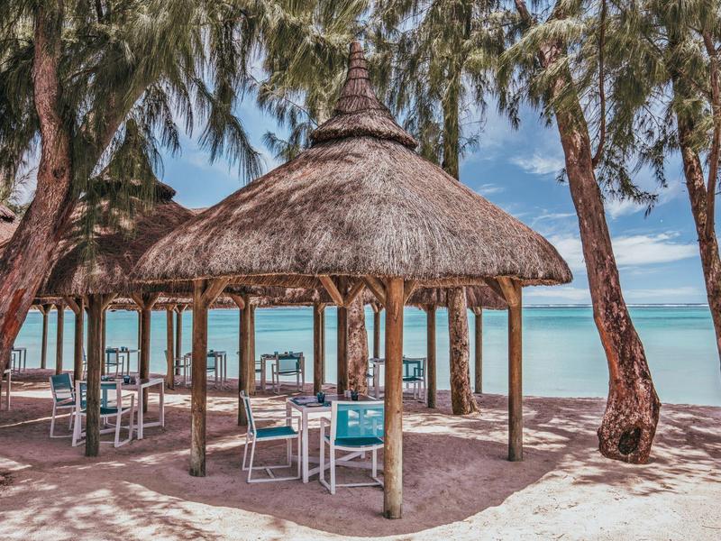 Strand mit Strohdächern, weißen Stühlen und Tischen unter Bäumen vor türkisfarbenem Meer.