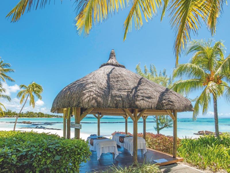 Offene Strohhütte mit Sitzplatz am weißen Sandstrand, umgeben von Palmen und blauem Himmel.