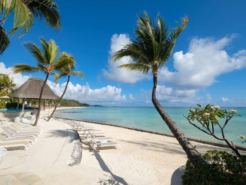 Strand mit weißem Sand, Palmen und Liegestühlen unter blauem Himmel mit vereinzelten Wolken.