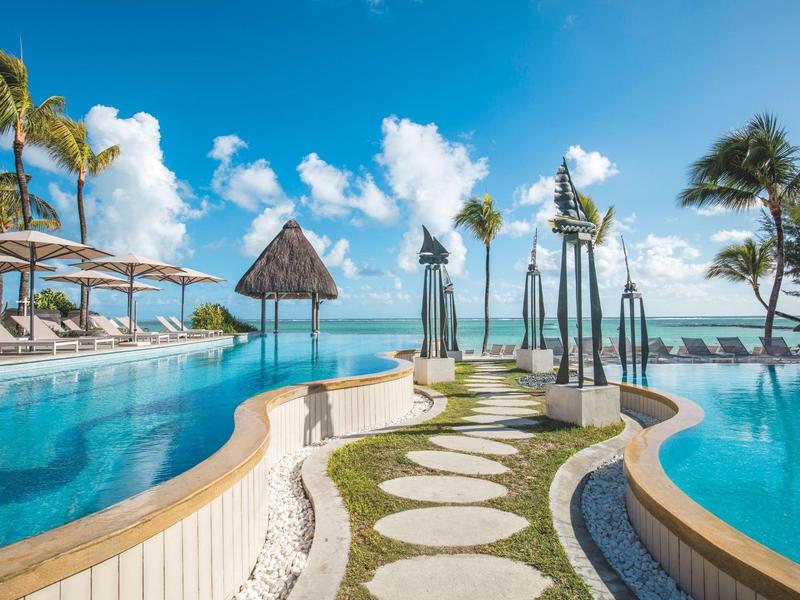 Luxuriöser Poolbereich mit Sonnenschirmen, Liegestühlen und Palmen unter blauem Himmel am Meer.