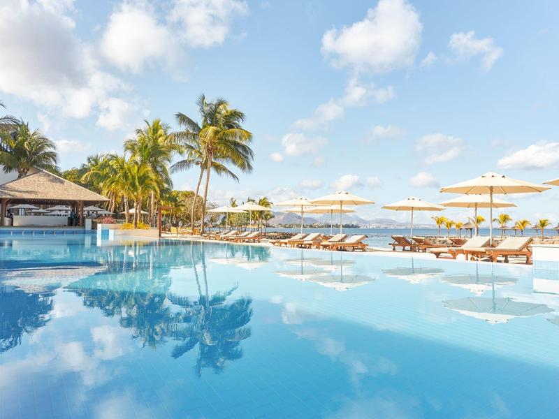Large pool with sun loungers and umbrellas beside palm trees under a blue sky.