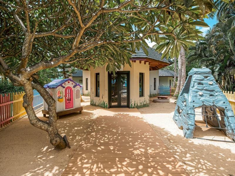 Children's playground with trees, sandy ground, and a small playhouse next to a hotel.