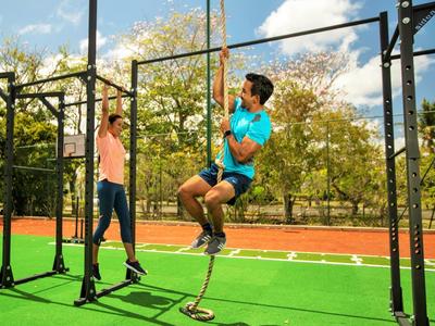 Zwei Personen trainieren draußen an einer Metall-Kletterstange auf grünem Sportplatz bei Sonnenschein.