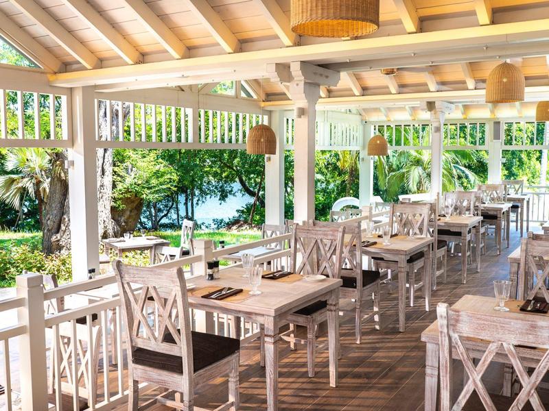 Licht, open terras met houten tafels en stoelen, omgeven door groen en uitzicht op water.