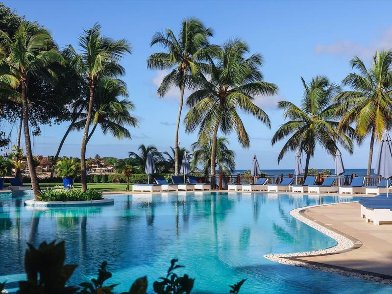 Groot zwembad met palmbomen en ligstoelen onder een blauwe hemel bij de zee