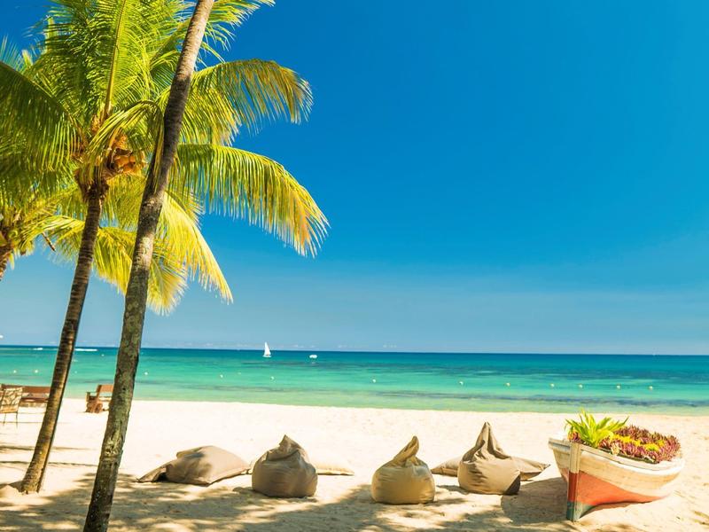 Strand met palmbomen, zand, zitzakken en een kleine boot onder een heldere blauwe lucht.
