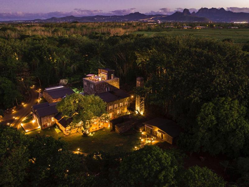 Een verlicht hotelgebouw omgeven door dicht bos met bergen op de achtergrond bij schemering.