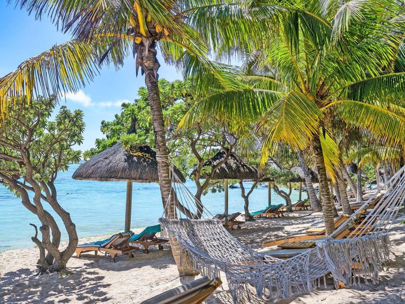 Strand mit Palmen, Hängematten und Strohdächern, türkisfarbenes Meer im Hintergrund.