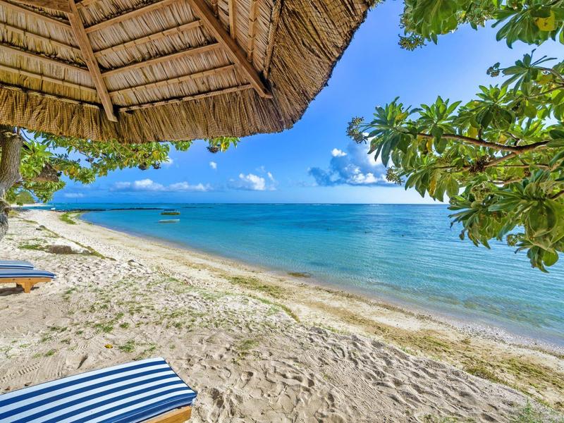Strand mit klarem blauen Wasser, weißem Sand, grünem Laub und Strohdach-Sonnenschirm.