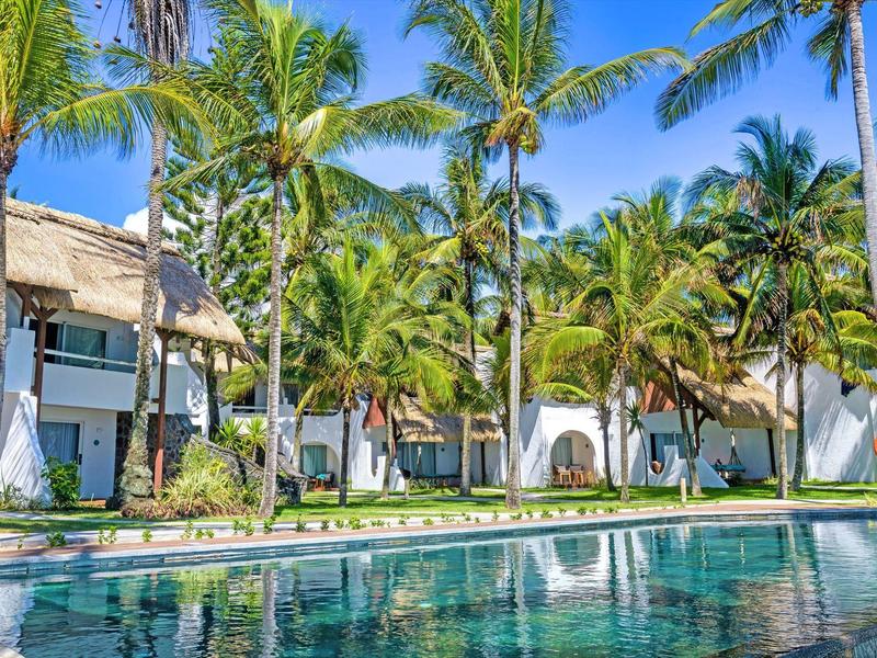 Einladender Pool vor weißen Bungalows, umgeben von hohen Palmen und blauem Himmel.