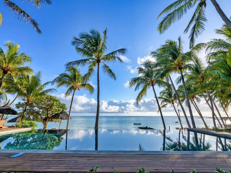 Infinity-Pool am Meer, umgeben von hohen Palmen und klarem blauem Himmel mit Wolken.