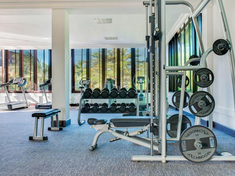 Licht fitnessruimte met gewichten, trainingsapparatuur en groen uitzicht buiten door ramen.