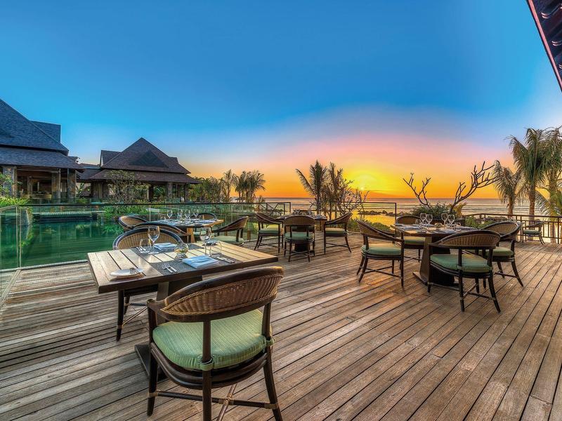 Houten loopbrug met glazen leuning leidt naar een modern hotel met uitzicht op zee bij zonsondergang.