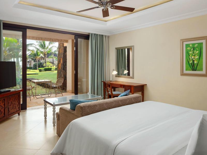 Hotelkamer met bed, bank, tv en uitzicht op een terras en tuin met palmbomen.