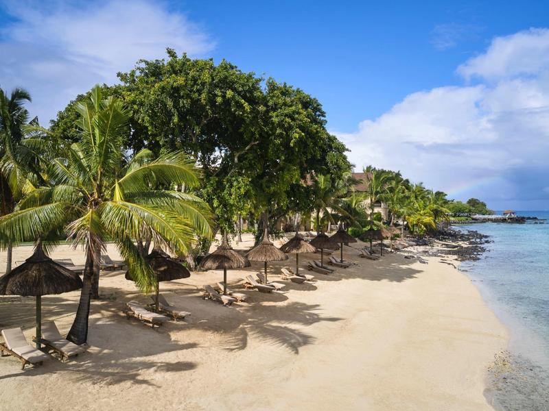 Een tropisch strand met palmbomen, parasols en helder blauw water onder een blauwe lucht.