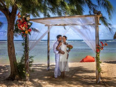 Paar bei einer Strandhochzeit unter weißem Baldachin mit Blumen, Meer im Hintergrund.