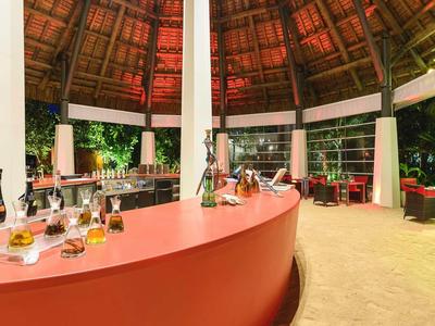 Otwarty bar z czerwonym kontuarem pod wysokim, okrągłym dachem ze strzechy w tropikalnym otoczeniu.