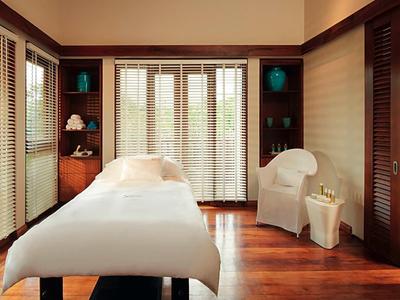 Jasny pokój zabiegowy z leżanką do masażu, drewnianą podłogą i dużymi oknami.