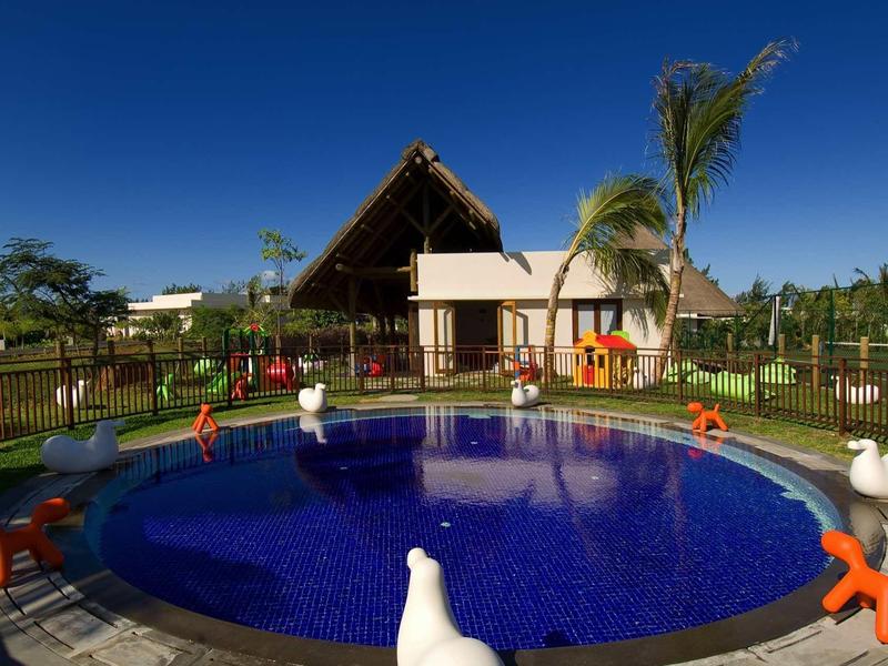 Okrągły basen z niebieskimi kafelkami i figurkami zwierząt przed budynkiem hotelu z dachem krytym strzechą