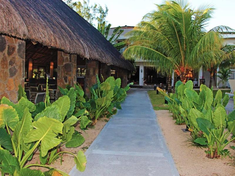Allée bordée de plantes tropicales menant à un bâtiment en chaume et des palmiers dans le jardin de l'hôtel.