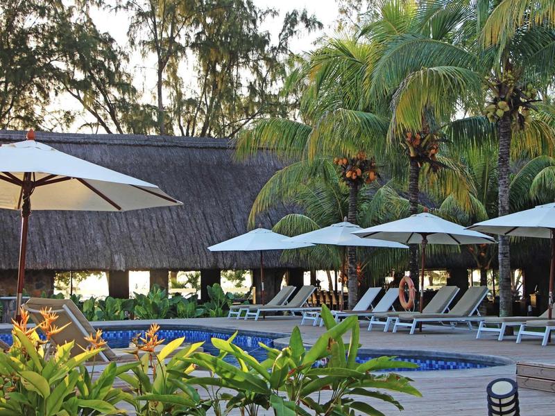Espace piscine avec transats et parasols entouré de palmiers et de plantes.