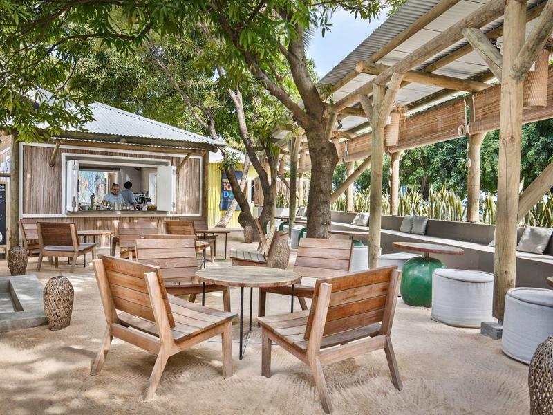 Wooden furniture and tables in a shaded, open-air lounge with a bar.