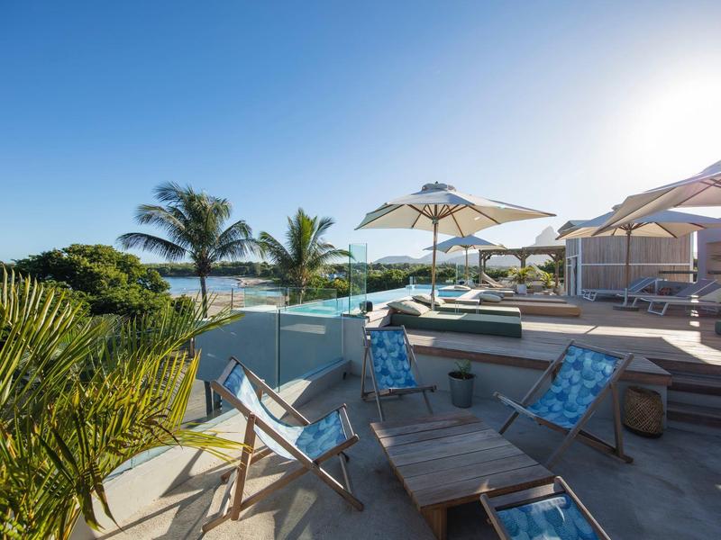 Sunny terrace with lounge chairs, umbrellas, and palm trees by the water.