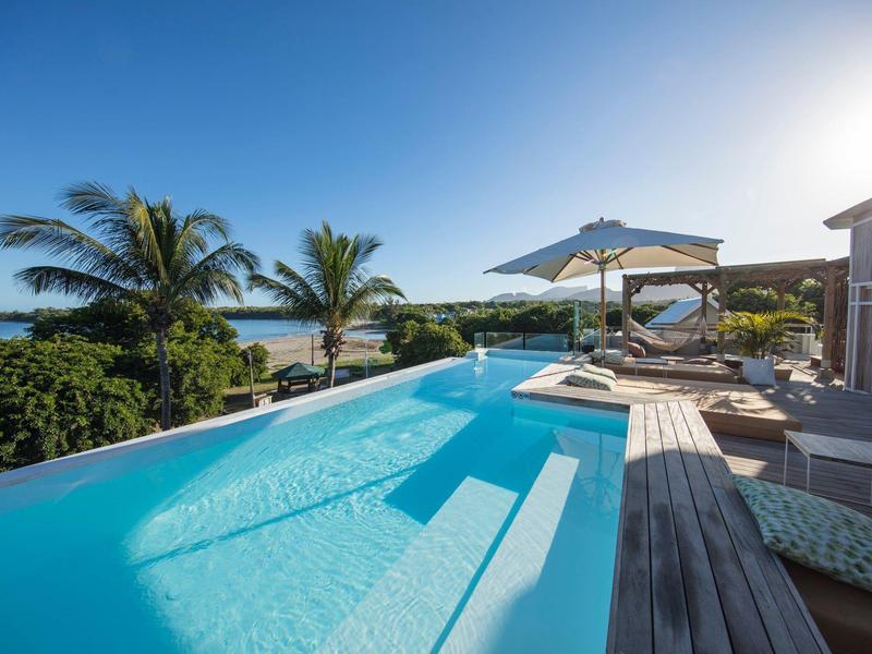 Infinity pool overlooking coast with palm trees and sun umbrella on wooden deck under clear sky