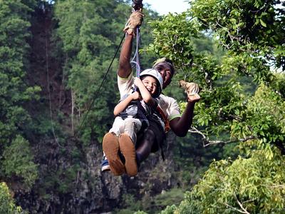 Zwei Personen beim Ziplining über grünen Bäumen, eine hält eine Getränkedose.