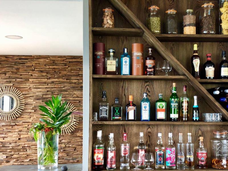 Holzregal mit Spirituosenflaschen, links Wand mit Steinoptik und Spiegel, Vase mit grünen Pflanzen.