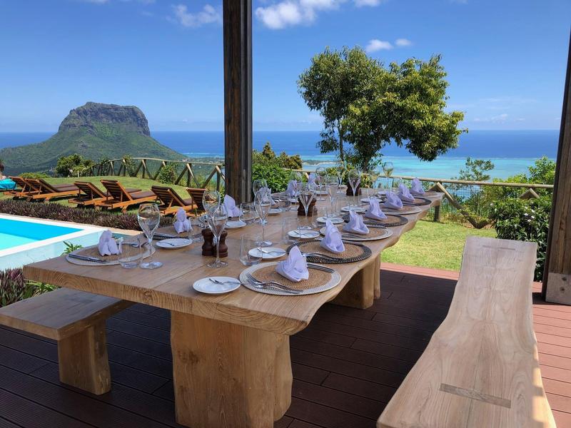 Langer Holztisch mit eingedeckten Tellern im Freien, mit Blick auf Meer und Insel im Hintergrund.